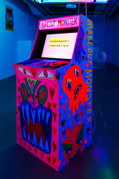 A colourfully painted arcade cabinet for the game Makeout Monster. It is in a colourfully lit room